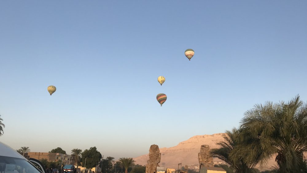 Four colorful hot air balloons float over Luxor, Egypt, with the Colossi of Memnon statues and desert cliffs visible below